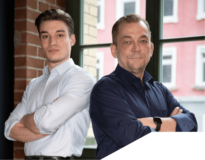 Jonas und Thomas Barnitzke stehen mit verschränkten Armen nebeneinander vor einem grossen Fenster mit Blick auf ein rotes Gebäude. Beide tragen Hemden. Sie schauen selbstbewusst in die Kamera.