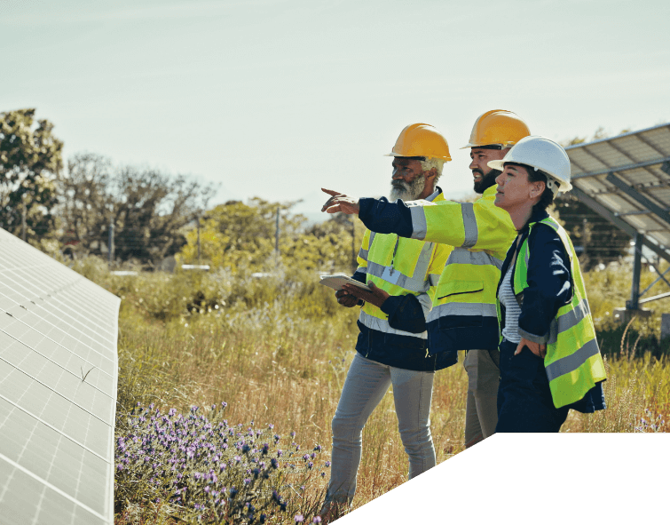 Drei Ingenieure oder Techniker in Sicherheitswesten und Helmen inspizieren eine Solaranlage im Freien. Einer von ihnen zeigt auf die Solarpanels, während die anderen aufmerksam zuhören. Die Anlage ist von grüner Vegetation umgeben.